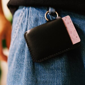 May include: A black leather wallet with a pink card holder attached to it. The wallet is hanging from a belt loop on a pair of blue jeans.