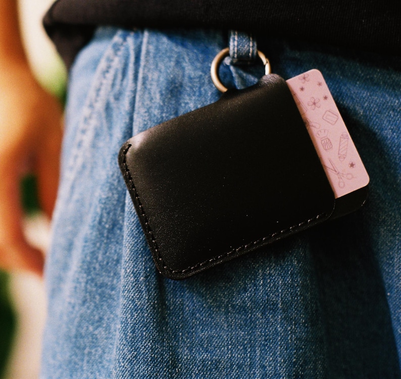 May include: A black leather wallet with a pink card holder attached to it. The wallet is hanging from a belt loop on a pair of blue jeans.