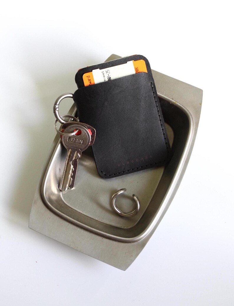 May include: A black leather wallet with a key ring and a key attached. The wallet is sitting in a silver metal dish.