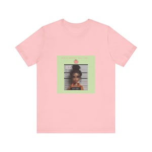 Puede incluir: Una camiseta de manga corta de color rosa claro con un gráfico central sobre un fondo cuadrado verde claro. El gráfico muestra una muñeca con un mono naranja frente a un fondo de ficha policial con el texto LEILA y APPLE DOESNT FALL FAR.