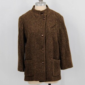 May include: A brown tweed jacket with a stand-up collar, three buttons, and two front pockets. The jacket features a small pocket on the left chest and three-quarter length sleeves. The fabric has a textured appearance.