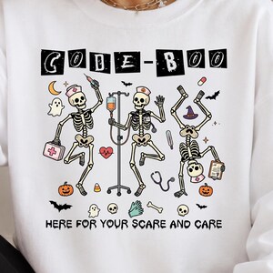 May include: A white long-sleeve sweatshirt featuring a graphic print of three skeletons dressed as medical staff. The text reads CODE-BOO at the top and HERE FOR YOUR SCARE AND CARE at the bottom, surrounded by small icons of pumpkins, ghosts, and bats.