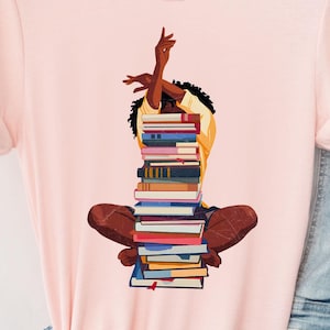 May include: A light pink short-sleeve t-shirt featuring a graphic illustration of a person with dark curly hair sitting cross-legged while balancing a tall stack of colorful books on their lap, with their arms raised above their head.