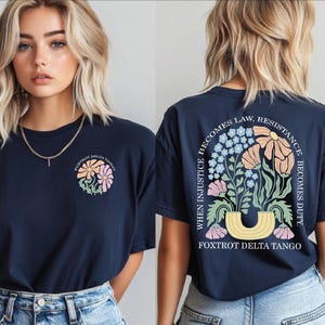 May include: Navy blue t-shirt with a floral design on the front and back. The front features a small floral graphic, while the back displays a larger arrangement with text that reads "When Injustice Becomes Law, Resistance Becomes Duty." The text "Foxtrot Delta Tango" is also present.