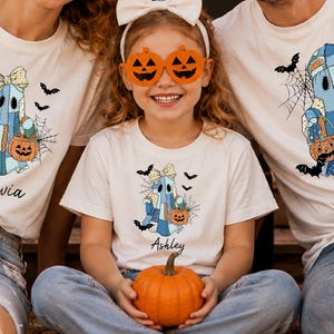 May include: Three white t-shirts featuring a graphic of a blue plaid ghost holding a pumpkin, with spiderwebs and bats. The shirts are personalized with the names Olivia, Ashley, and Alex. The individuals wear orange pumpkin-shaped glasses.