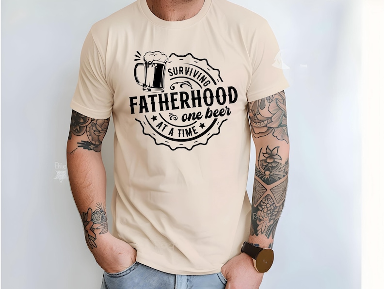 May include: A beige t-shirt with a black graphic that says "Surviving Fatherhood one beer at a time". The graphic includes a beer mug with a foam head.