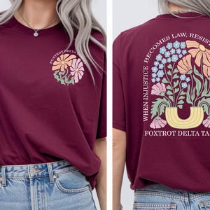 May include: Burgundy t-shirt with a floral design on the front and back. The front features a flower graphic with the text "Foxtrot Delta Tango." The back displays a floral arrangement with the text "When injustice becomes law, resistance becomes duty."
