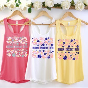 May include: Three tank tops in coral, white, and yellow hang on wooden hangers. Each top features a floral design with the words "Freedom Democracy Truth" or "Freedom Democracy Ruth". The tops are decorated with white, pink, and blue flowers.