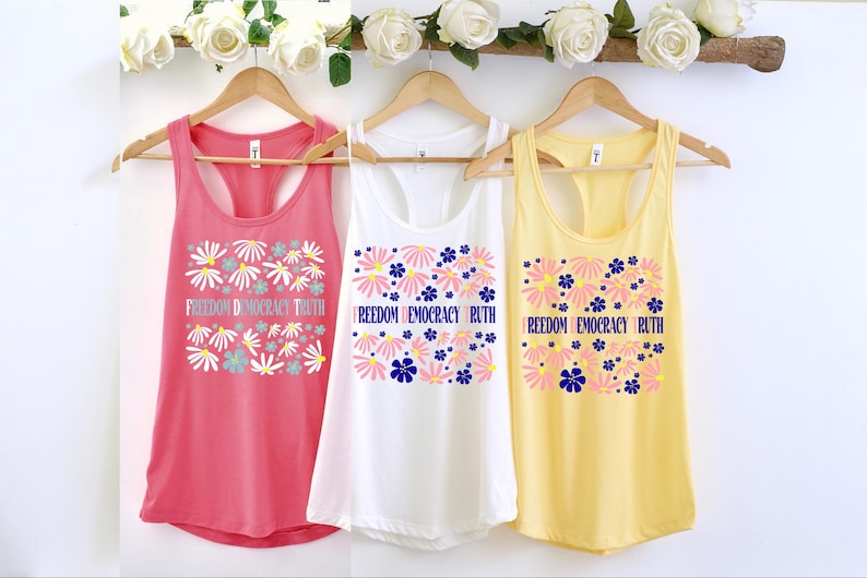 May include: Three tank tops in coral, white, and yellow hang on wooden hangers. Each top features a floral design with the words "Freedom Democracy Truth" or "Freedom Democracy Ruth". The tops are decorated with white, pink, and blue flowers.