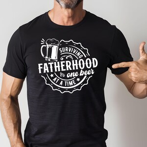May include: Black t-shirt with a white graphic design of a beer mug and the text "Surviving Fatherhood one beer at a time".