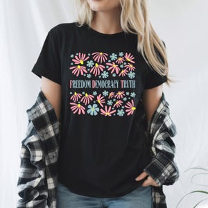 May include: Black t-shirt with a floral design in pink, blue, and yellow. The text "FREEDOM DEMOCRACY TRUTH" is printed below the flowers. The shirt is a crew neck and short-sleeved.
