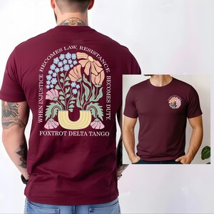 May include: Burgundy t-shirt with a floral design on the back and a smaller version on the front. The back design features flowers, leaves, and the text "WHEN INJUSTICE BECOMES LAW, RESISTANCE BECOMES DUTY. FOXTROT DELTA TANGO."