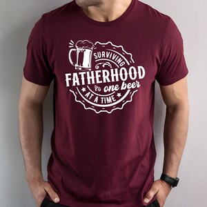 May include: Burgundy t-shirt with a white graphic of a beer mug and the text "Surviving Fatherhood one beer at a time".