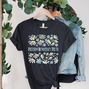 May include: Dark gray t-shirt with a floral design featuring white and light blue flowers with yellow centers. The text "FREEDOM DEMOCRACY TRUTH" is centered below the flowers. The shirt is hanging on a wooden hanger.