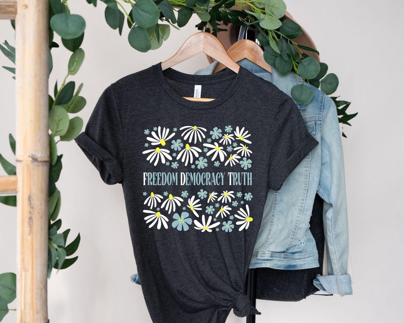 May include: Dark gray t-shirt with a floral design featuring white and light blue flowers with yellow centers. The text "FREEDOM DEMOCRACY TRUTH" is centered below the flowers. The shirt is hanging on a wooden hanger.