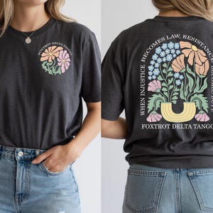 May include: Dark gray t-shirt with a floral design on the front and back. The front features a cluster of pastel flowers and the text "FOXTROT DELTA TANGO." The back displays a floral arrangement with the text "WHEN INJUSTICE BECOMES LAW, RESISTANCE BECOMES DUTY."