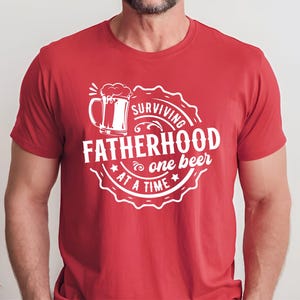May include: Red t-shirt with a white graphic design of a beer mug and the text "Surviving Fatherhood one beer at a time".