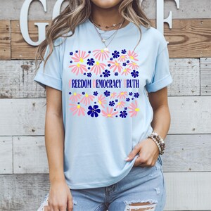 May include: Light blue t-shirt with a floral design. The design includes pink and blue flowers with the words "FREEDOM DEMOCRACY TRUTH" in a light purple color. The shirt is made of a soft material.