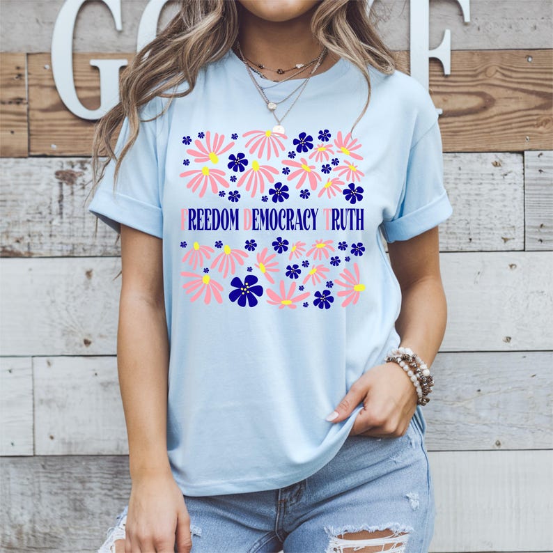 May include: Light blue t-shirt with a floral design. The design includes pink and blue flowers with the words "FREEDOM DEMOCRACY TRUTH" in a light purple color. The shirt is made of a soft material.