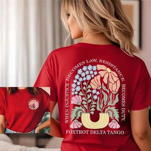 May include: A red t-shirt with a floral design on the back, featuring a variety of flowers in pastel colors and the text "WHEN INJUSTICE BECOMES LAW, RESISTANCE BECOMES DUTY." The shirt also has a smaller floral design on the front.