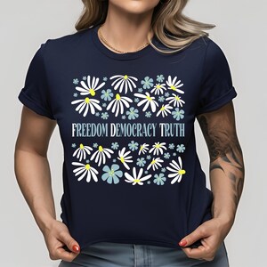 May include: Navy blue t-shirt with a floral design and the words "FREEDOM DEMOCRACY TRUTH" in light blue. The floral design features white and light blue flowers with yellow centers.