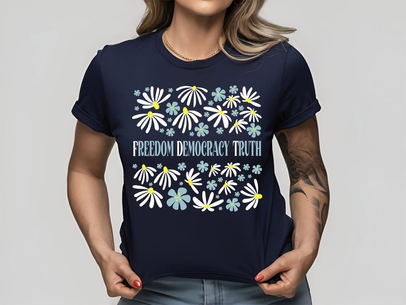 May include: Navy blue t-shirt with a floral design and the words "FREEDOM DEMOCRACY TRUTH" in light blue. The floral design features white and light blue flowers with yellow centers.
