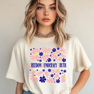 May include: Cream-colored t-shirt featuring a floral design with pink and blue flowers. The words "FREEDOM DEMOCRACY TRUTH" are printed in blue across the center of the shirt. The shirt has short sleeves.