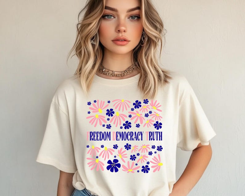 May include: Cream-colored t-shirt featuring a floral design with pink and blue flowers. The words "FREEDOM DEMOCRACY TRUTH" are printed in blue across the center of the shirt. The shirt has short sleeves.