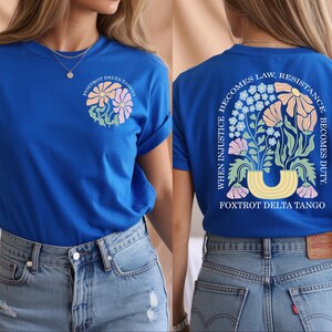 May include: Royal blue t-shirt with a floral design on the front and back. The front features a circular floral design with the text "Foxtrot Delta Tango". The back has a floral arch with the text "When injustice becomes law, resistance becomes duty."