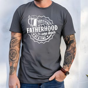 May include: A dark grey t-shirt with a white graphic design that says "Surviving Fatherhood one beer at a time". The graphic features a beer mug with a foam head.