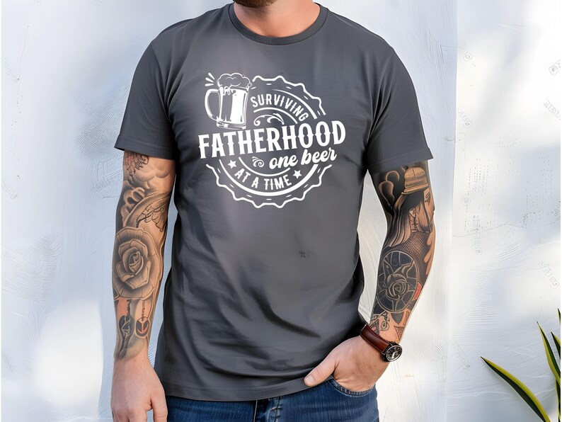May include: A dark grey t-shirt with a white graphic design that says "Surviving Fatherhood one beer at a time". The graphic features a beer mug with a foam head.