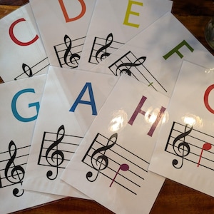 Puede incluir: Un conjunto de tarjetas de memoria con las letras C a G, cada una con un color diferente y una nota musical en un pentagrama. Las notas son C, D, E, F, G, A y H.