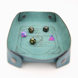 May include: A teal leather dice tray with a black and gold dice set. The tray has a design that includes the words "Difficulty Class 20" and "All Dice".