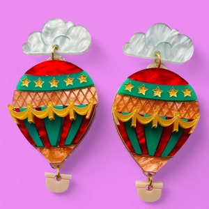 May include: A pair of red, green, and gold hot air balloon earrings with a white cloud charm. The earrings have a gold hook and a white plastic backing.