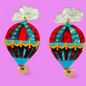 May include: A pair of red, blue, and gold hot air balloon earrings with white cloud accents. The earrings are made of acrylic and have a whimsical design.