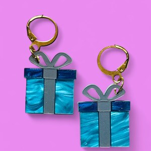 May include: A pair of earrings shaped like gift boxes. The boxes are turquoise with a marbled effect, accented with a dark blue top and a silver ribbon. The earrings have gold-colored leverback closures.
