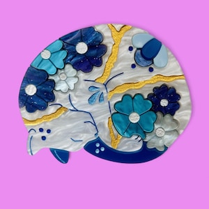 May include: A white ceramic brooch shaped like a sleeping cat with blue and gold floral details.