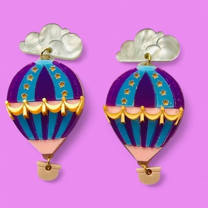 May include: A pair of purple, blue, and gold hot air balloon earrings with white cloud accents. The earrings are made of resin and have a whimsical design.