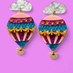 May include: A pair of hot air balloon earrings with pink, purple, blue, and gold accents. Each earring features a white cloud charm.