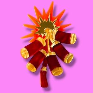 Op de afbeelding: Decoratieve broche met een cluster van rode cilinders met gouden accenten, die op vuurwerk lijken. Het ontwerp omvat een oranje uitbarsting en een gouden centrum, tegen een roze achtergrond.