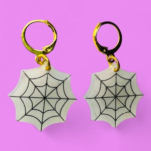 Puede incluir: Un par de pendientes de aro dorados con dijes de telaraña blanca. Los dijes tienen forma de telaraña con líneas negras.