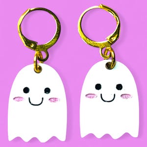 Pode incluir: Um par de brincos de argola dourados com pingentes de fantasmas brancos. Cada fantasma tem um rosto sorridente preto e bochechas rosadas.