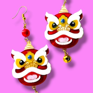 Op de afbeelding: Een paar rood, wit en goudkleurige oorbellen met leeuwendans. Elke oorbel heeft een leeuwenkop met een rode body, witte manen en gouden accenten. Een kleine gouden bel hangt aan de onderkant, en een rode kraal zit bovenaan.