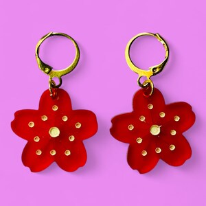 Pode incluir: Um par de brincos vermelhos em forma de flor com argolas douradas. Os brincos têm um centro dourado e pontos brancos ao redor do centro.