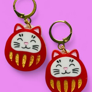 Pode incluir: Um par de brincos de gato vermelhos e dourados com um rosto sorridente. Os brincos são feitos de resina e têm um fecho de gancho dourado.