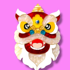 Op de afbeelding: Een kleurrijke leeuwendanshoofdbroche met een rood, geel, wit en goud ontwerp. De broche heeft een lachend gezicht met grote ogen en een gedetailleerde manen, tegen een roze achtergrond.