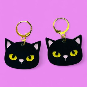 Pode incluir: Um par de brincos de rosto de gato preto com argolas douradas. Os rostos de gato têm olhos amarelos e narizes cor de rosa.