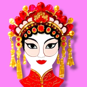Op de afbeelding: Decoratieve broche met een gestileerd gezicht van een vrouw in traditionele Chinese opera-outfit. De broche heeft een rode en gouden hoofdtooi, een rode jurk en een wit gezicht met zwarte ogen en rode lippen. De achtergrond is effen roze.