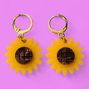 Pode incluir: Um par de brincos de argola dourados com pingentes de girassol amarelo. Os pingentes de girassol têm um centro castanho com um padrão de grelha.