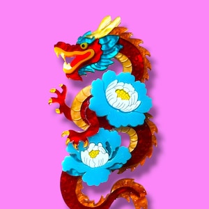 Op de afbeelding: Een kleurrijk drakenbeeldje met een rode body, gele accenten en blauwe bloemversieringen. De draak heeft een rode kop met gele hoorns en staat tegen een roze achtergrond. Het lichaam van de draak is versierd met twee blauwe bloemen.